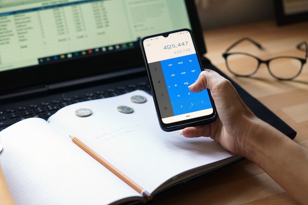 A person's hands using a mobile banking app on a smartphone, showing various savings account options with attractive interest rates. The background includes a calendar and financial charts.