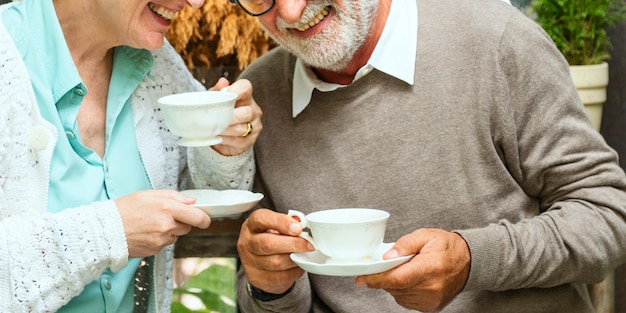 A close-up of an elderly couple smiling and holding mugs on a porch, with a tablet showing stock charts in the background. The scene suggests retirement income and financial security provided by dividend investments.