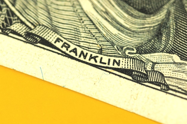 A close-up shot of the U.S. dollar bill with a blurred background showing various economic symbols, such as charts, graphs, and miniature buildings, representing the complexity of the financial market.
