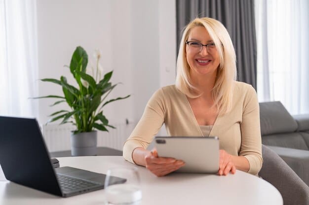 A middle-aged woman sitting at her home office desk, looking at a tablet displaying financial data and charts while smiling. Her hands are gently resting on the tablet screen.
